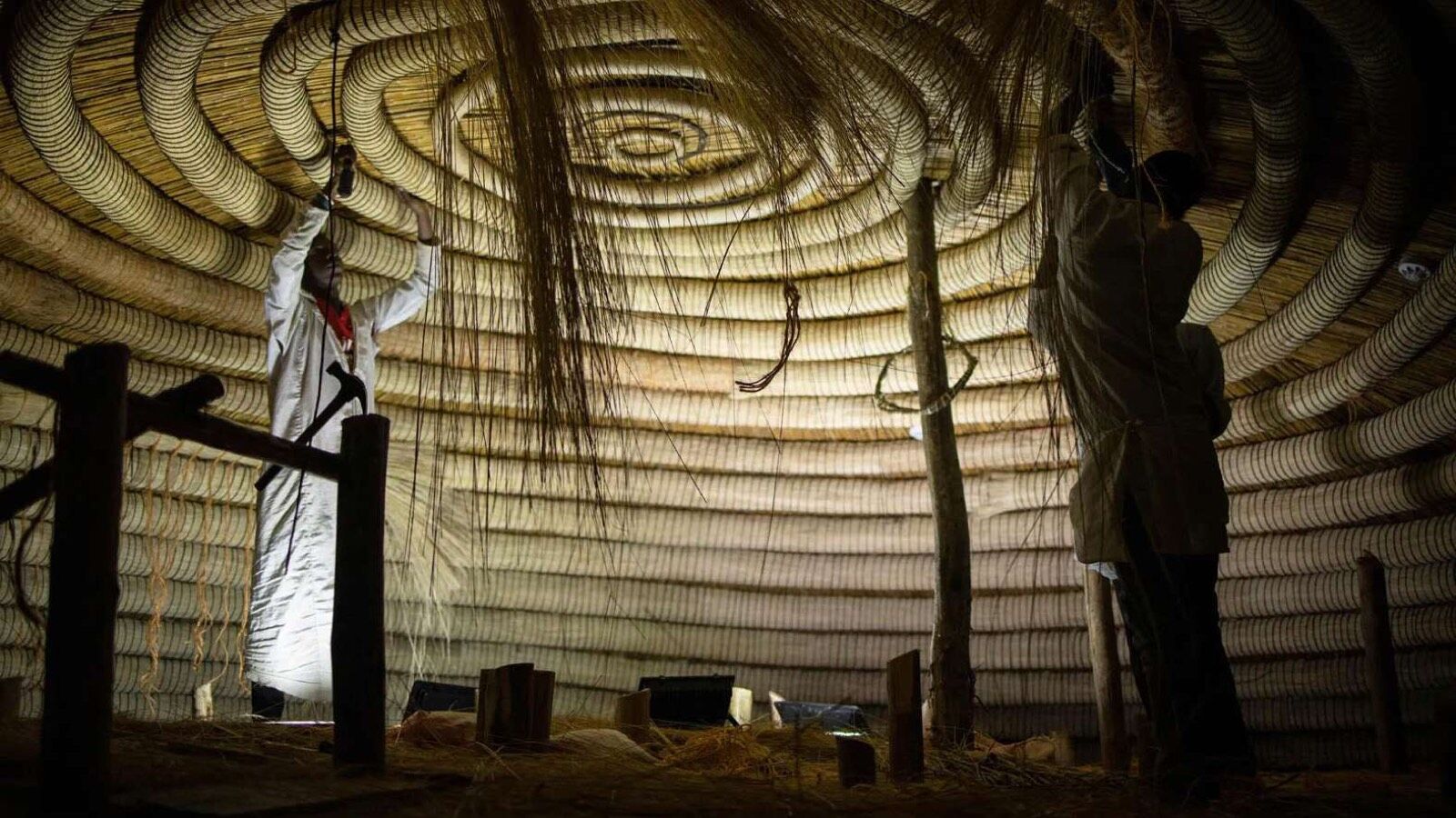 A weaver at work inside a thatched roof, the reed spiral above them
